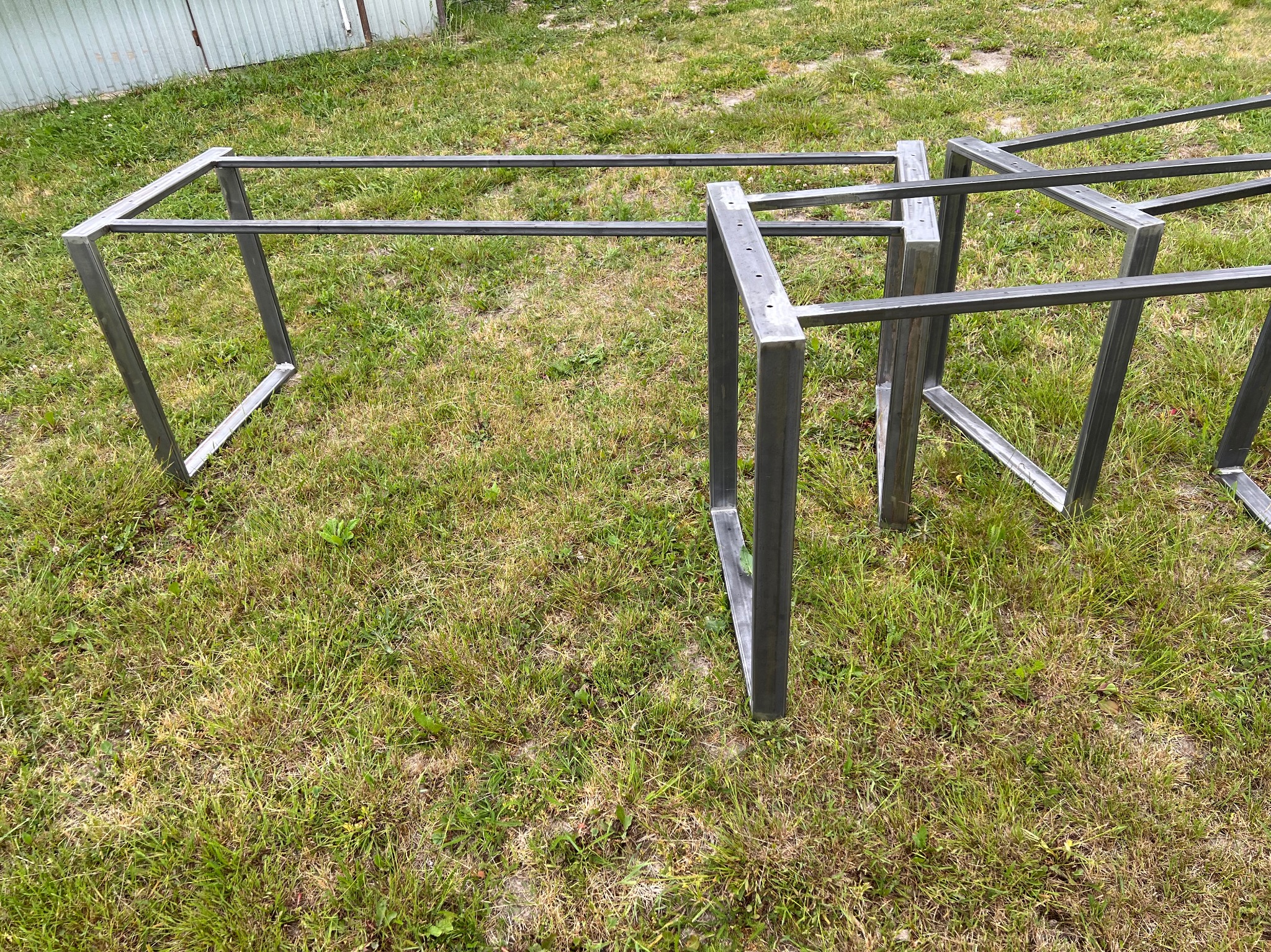 Surowe, spawane metalowe ramy stołów z otworami na blaty, rozłożone na trawie przed blaszakiem.