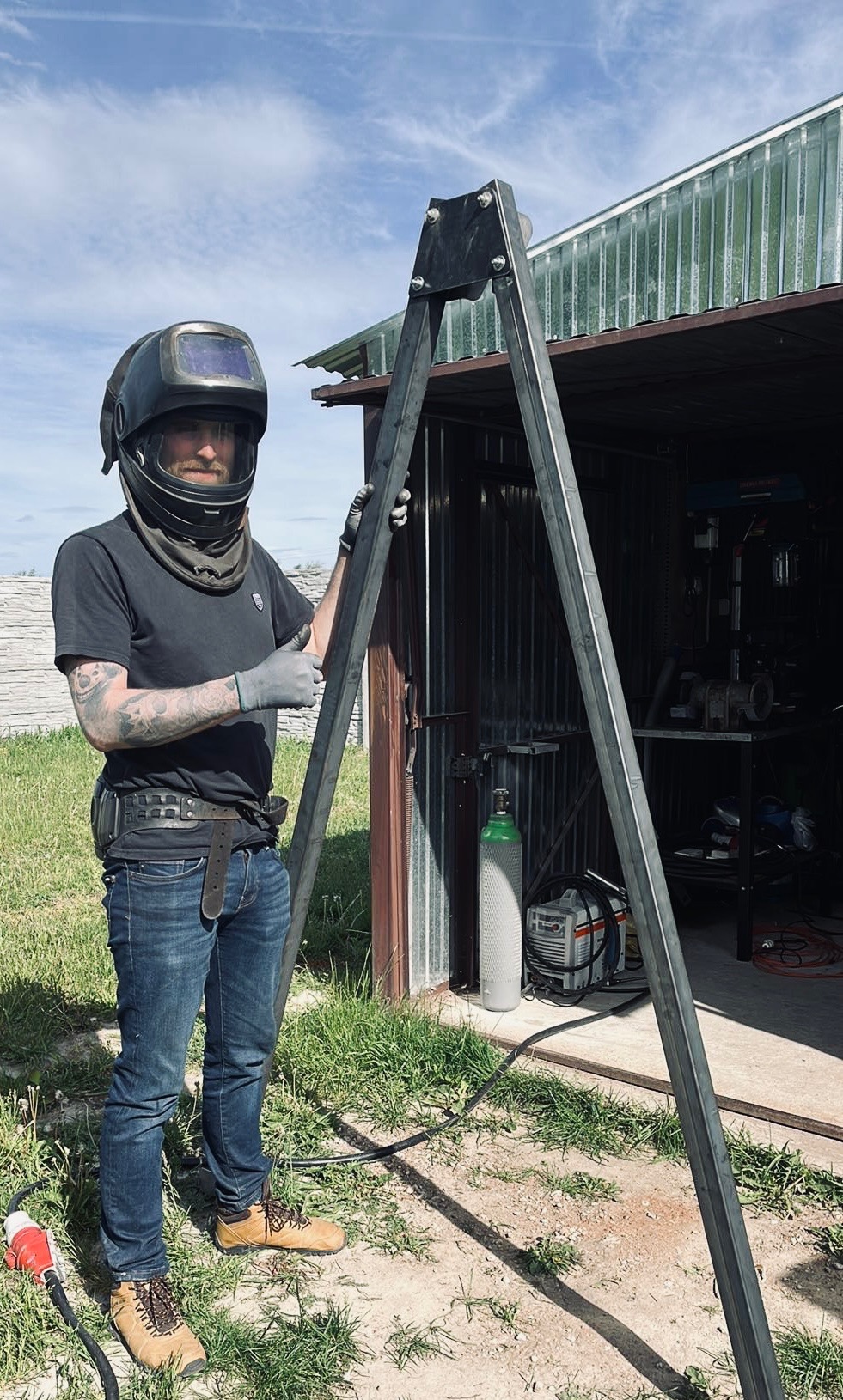 Spawacz w masce i rękawicach, prezentuje metalowy statyw spawalniczy na tle otwartego garażu z butlą gazową i spawarką, uniesiony kciuk w górę.