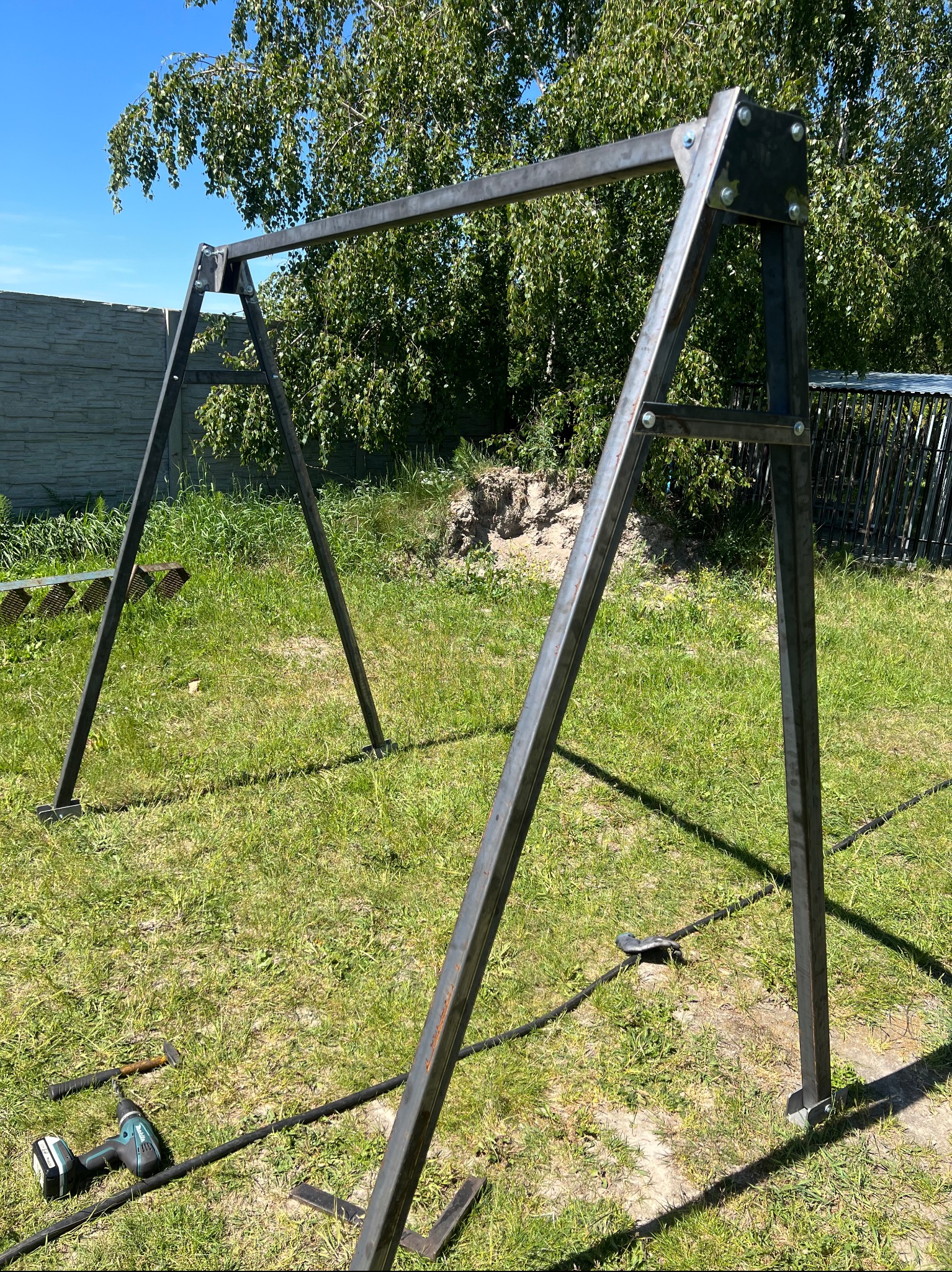 Surowa, metalowa konstrukcja huśtawki ogrodowej w trakcie montażu na trawniku, widoczne narzędzia i elementy mocujące.