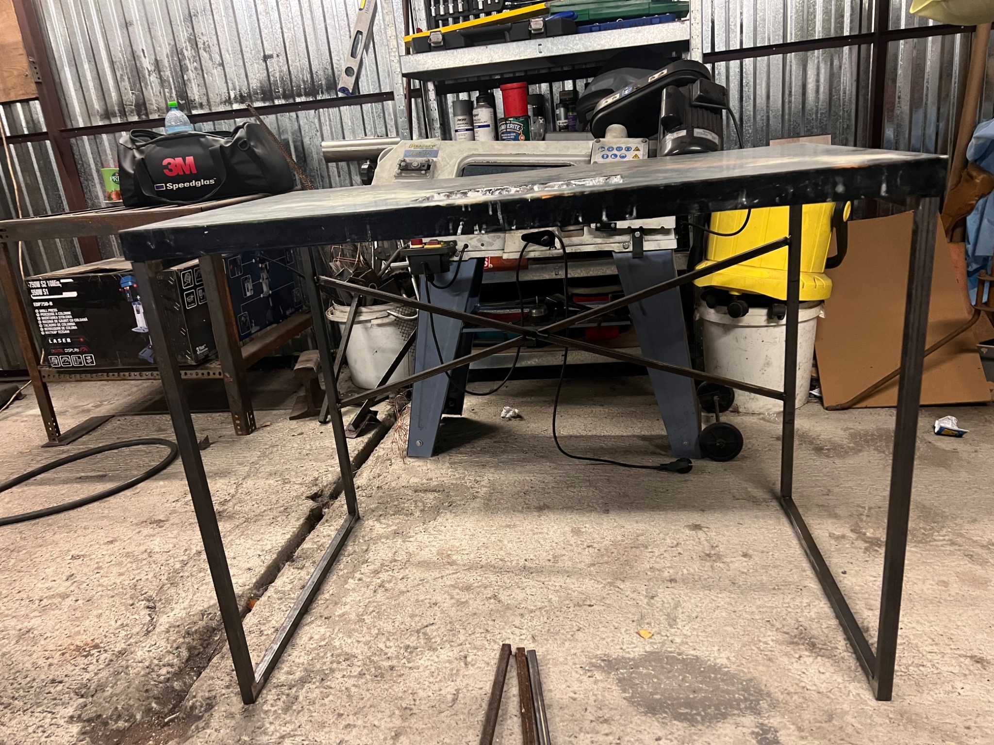 Stół roboczy z metalowych profili w trakcie spawania, widoczne ślady spawania na blacie, w tle warsztat z narzędziami i akcesoriami spawalniczymi.