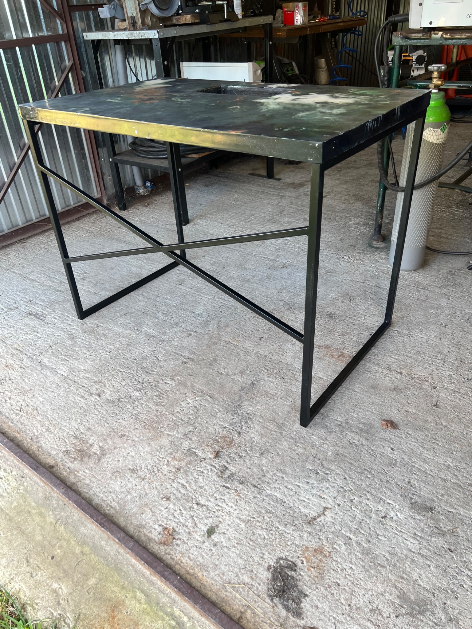 Stół z metalowym szkieletem i blatem o abstrakcyjnym wzorze, stojący w warsztacie, widoczny sprzęt spawalniczy w tle.