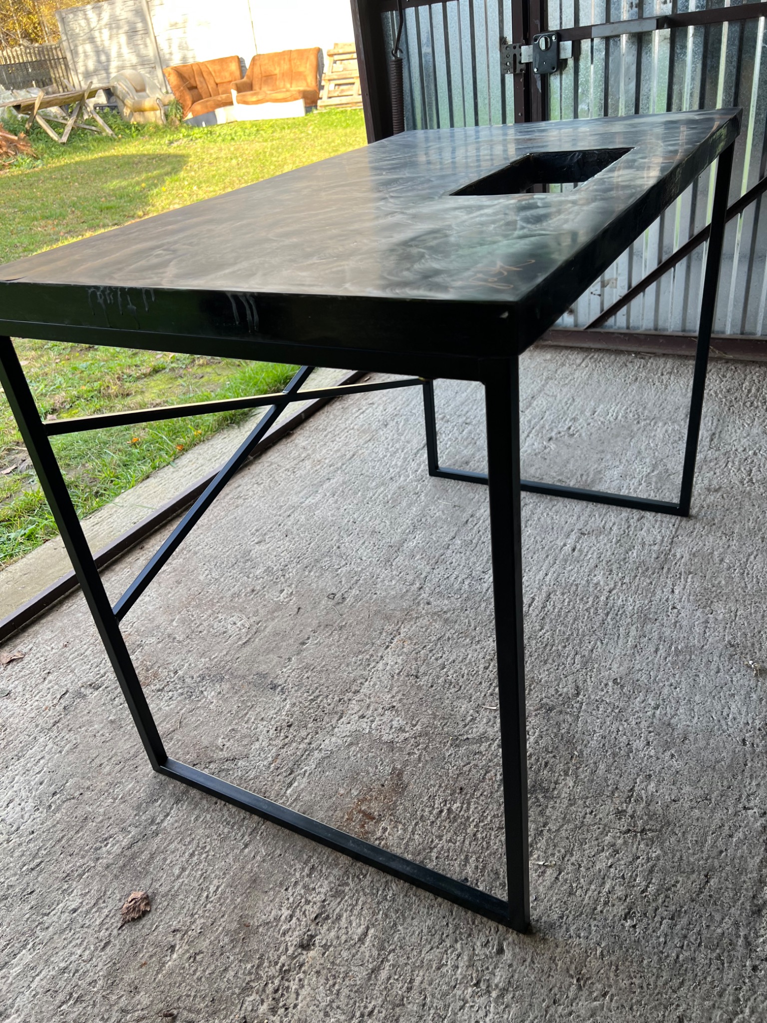 Stół metalowy z blatem imitującym marmur, czarny stelaż z profili kwadratowych, widoczny otwór w blacie, tło stanowią blaszane drzwi garażowe i fragment ogrodu z meblami ogrodowymi.