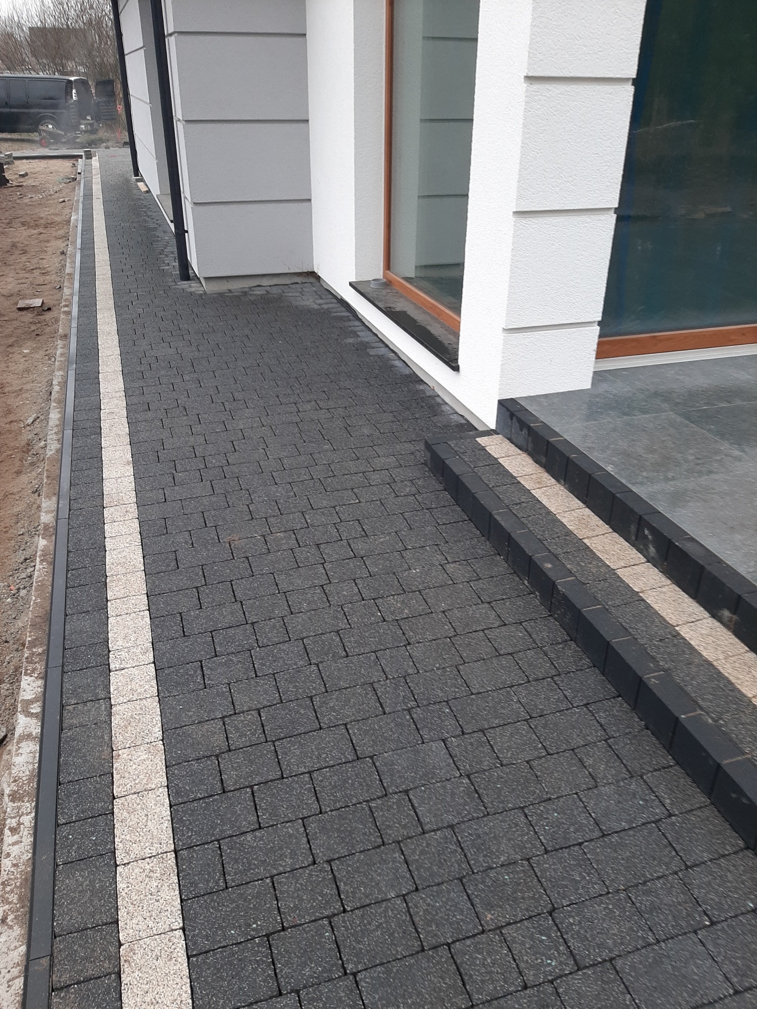 Ułożona kostka brukowa w kolorze grafitowym z jasnym pasem dekoracyjnym prowadząca do wejścia domu z widocznym oknem.