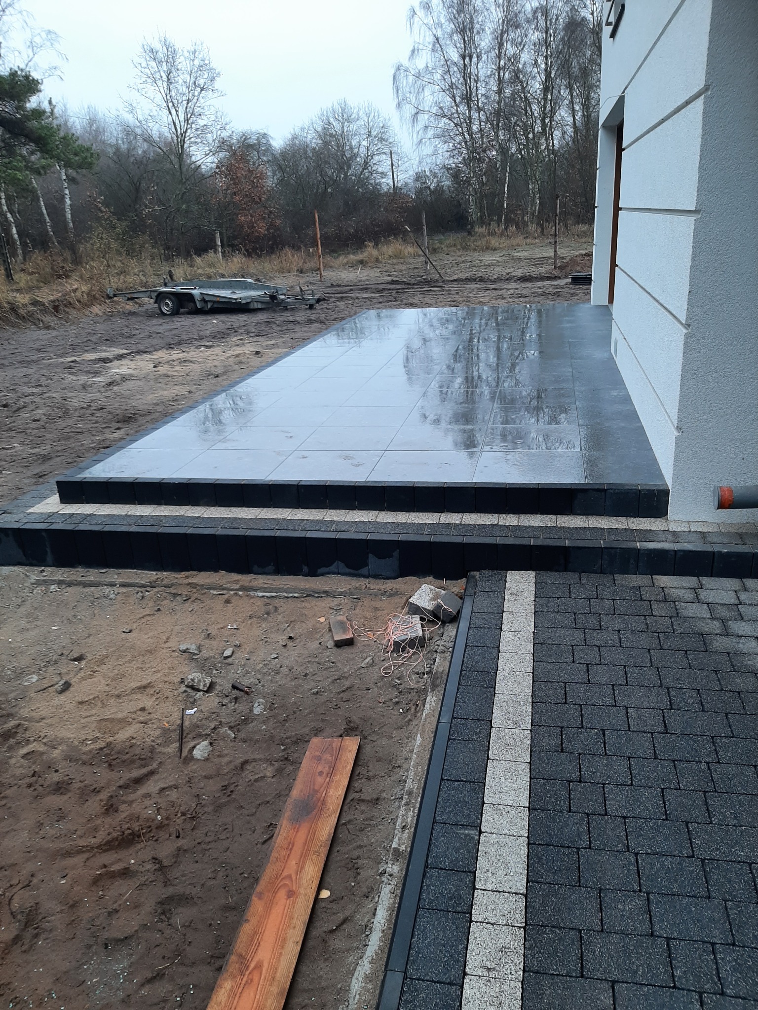 Nowo ułożony taras z szarych płytek ceramicznych przed domem, z widocznymi refleksami na mokrej powierzchni, obok ułożona kostka brukowa w kolorze grafitowym z jasnym pasem, w tle widoczna przyczepa.