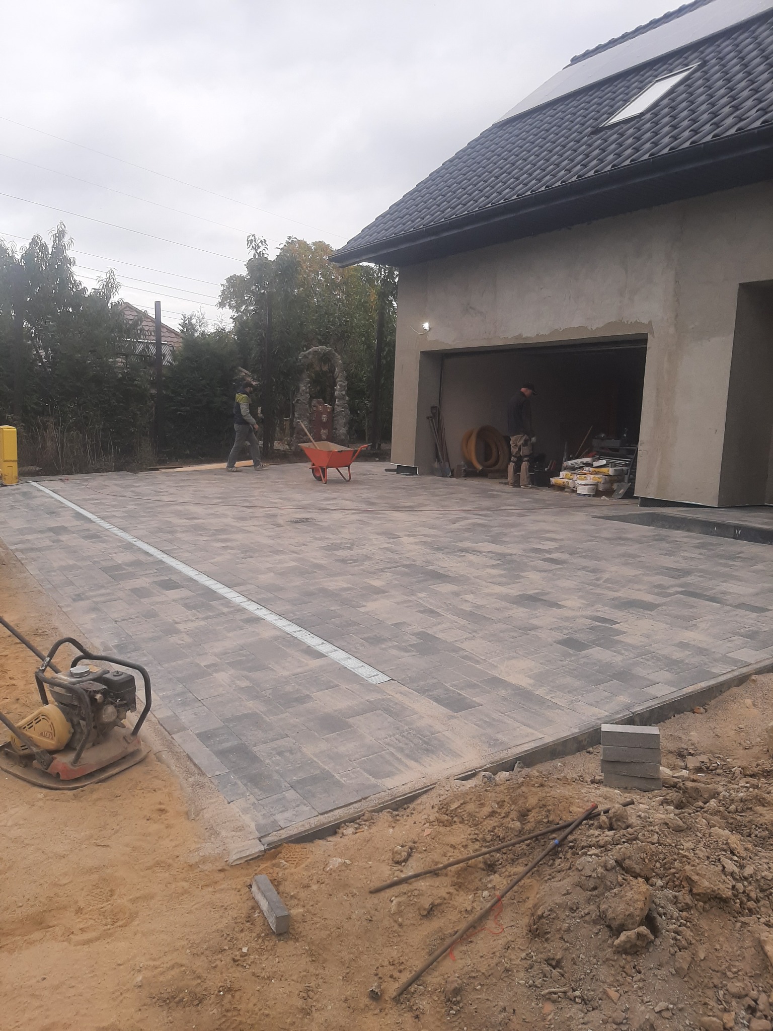 Ułożona kostka brukowa na podjeździe do garażu, widoczna ubijarka wibracyjna, pracownicy w tle i przygotowane materiały budowlane w garażu.