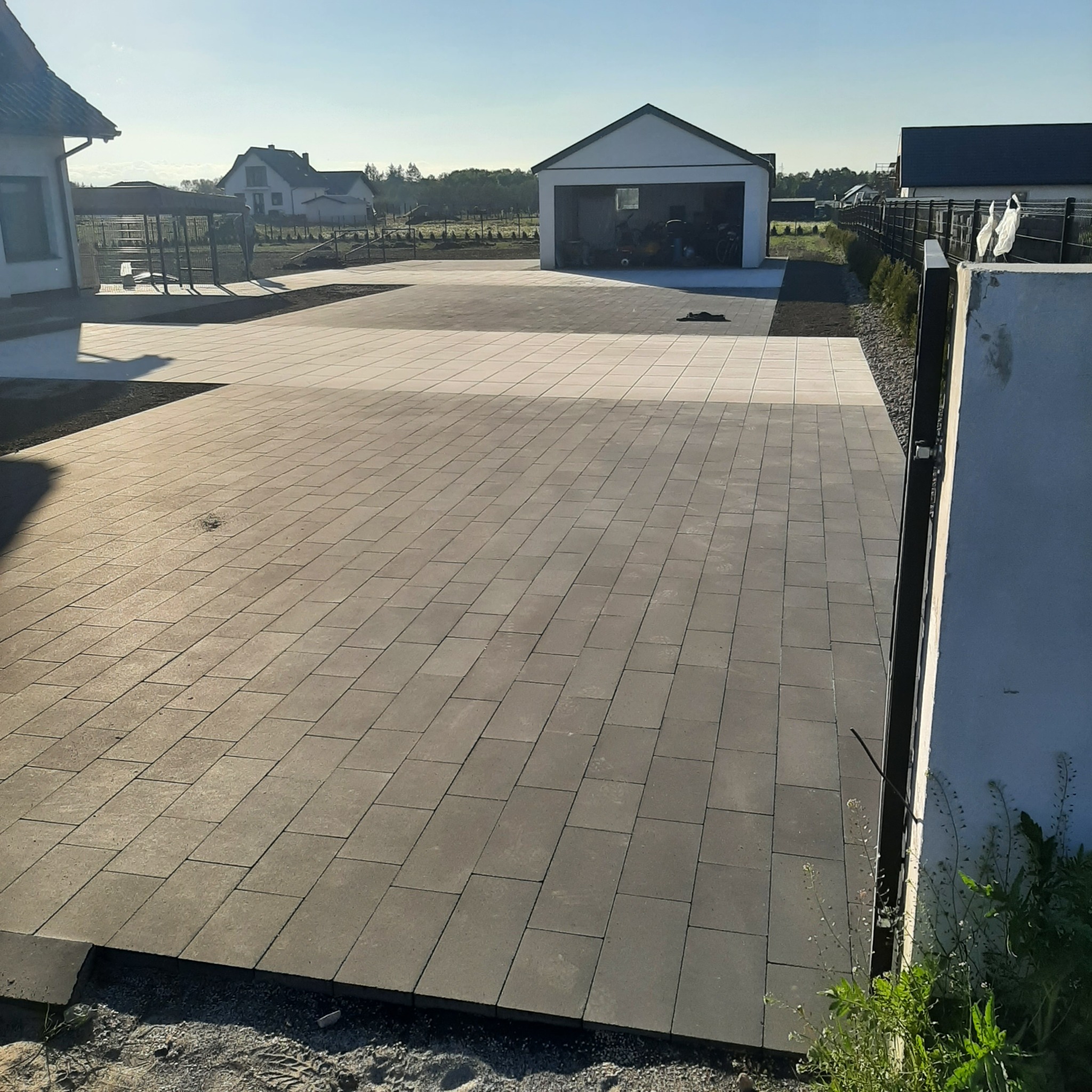 Ułożona, szara kostka brukowa na podjeździe przed domem i garażem w słoneczny dzień, widoczny fragment ogrodzenia i roślinność.