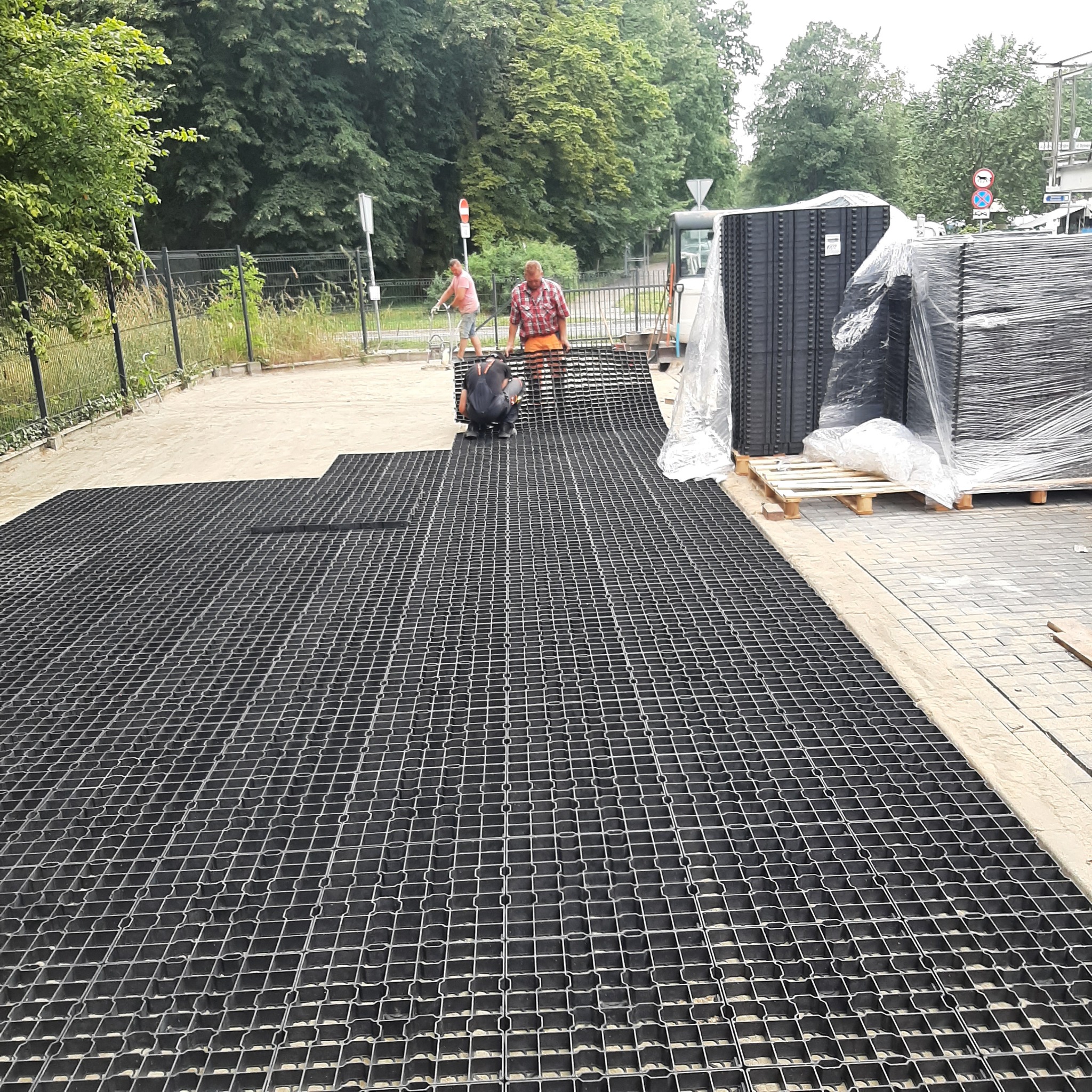 Montaż ażurowych płyt parkingowych na piaskowym podłożu przez dwóch pracowników, palety z płytami w tle, widoczne drzewa i ogrodzenie.