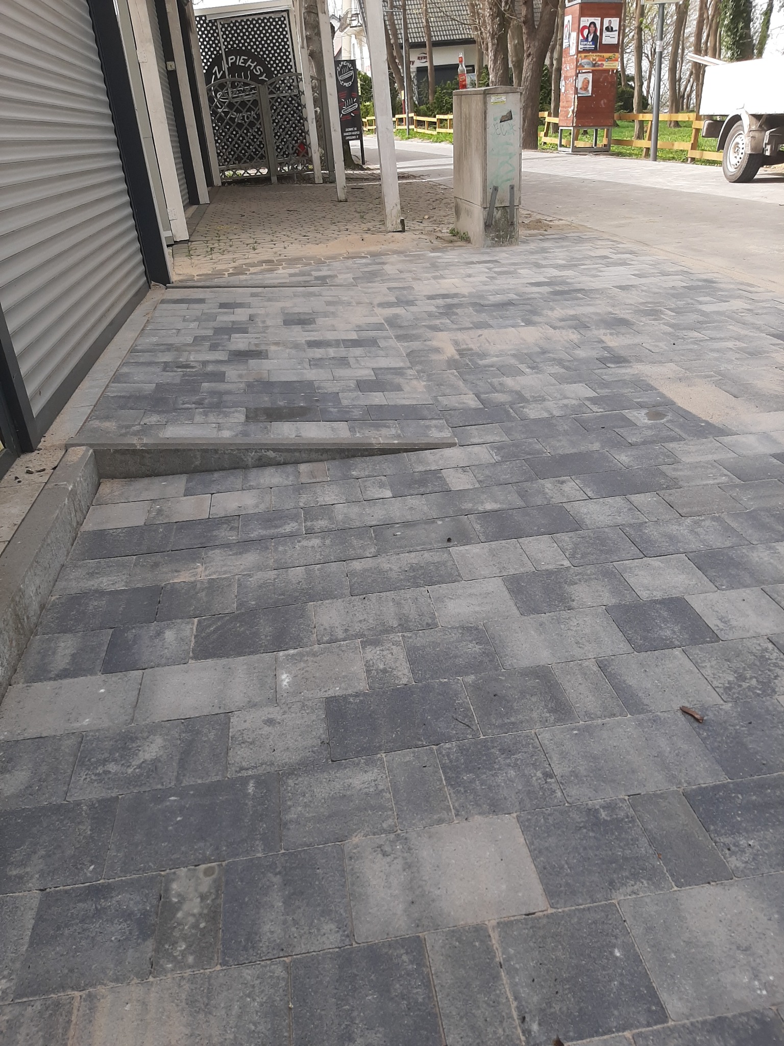 Ułożona szara kostka brukowa przed budynkiem z metalową roletą i słupami.