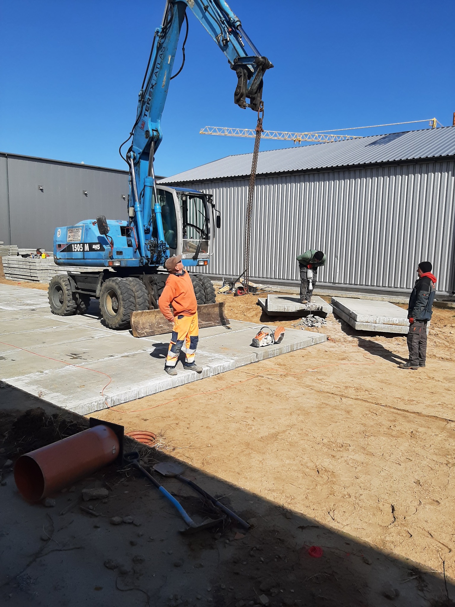 Niebieska koparka podnosi betonową płytę na placu budowy, w tle pracownicy z młotem pneumatycznym i piłą do betonu.