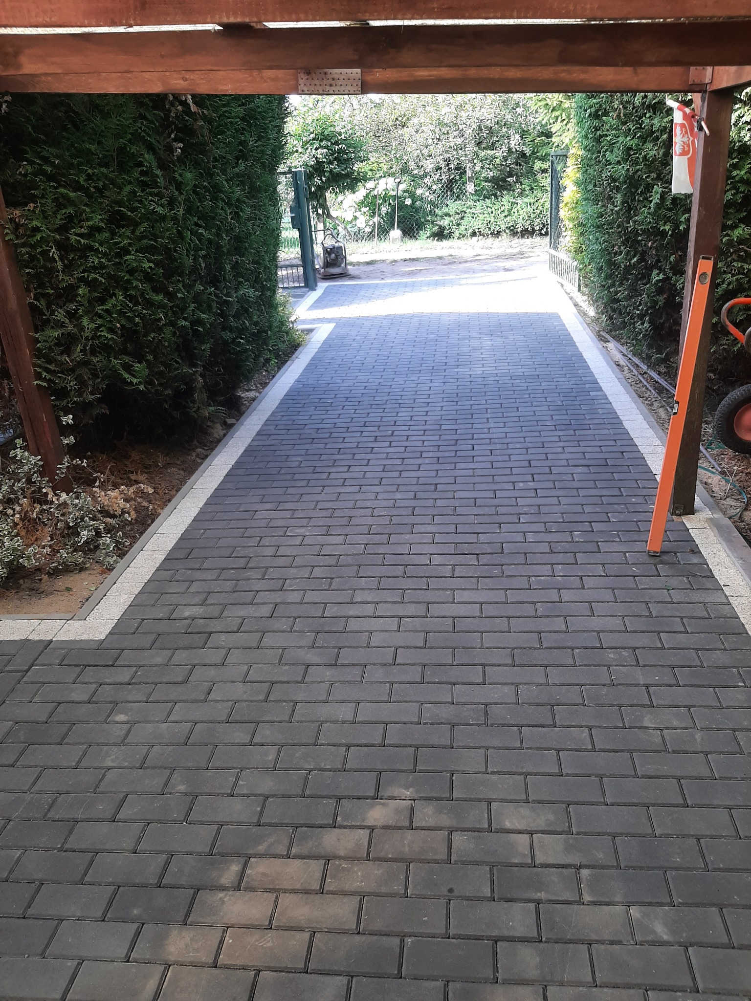 Ułożona ścieżka z ciemnoszarej kostki brukowej z jasnoszarym obramowaniem, prowadząca przez drewnianą konstrukcję w kierunku zielonego ogrodu, z widoczną poziomicą opartą o drewniany słup.