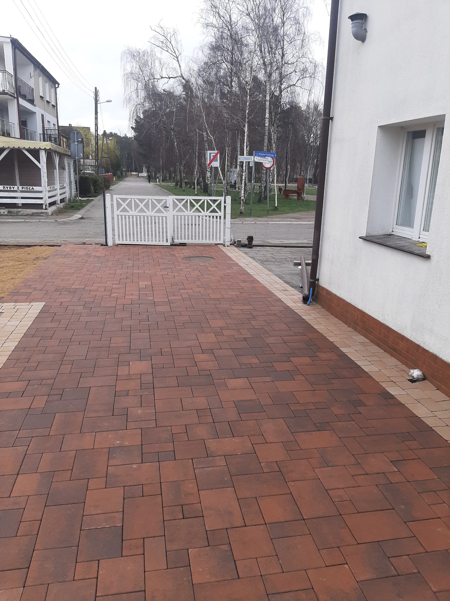 Nowo ułożona, czerwono-brązowa kostka brukowa na podjeździe, z białym ogrodzeniem w tle i fragmentem budynku po prawej stronie.