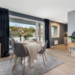 Jasne drewniane podłogi w nowoczesnym salonie z okrągłym stołem nakrytym na cztery osoby, szary dywan, ciemne zasłony i balkon z widokiem na zieleń.