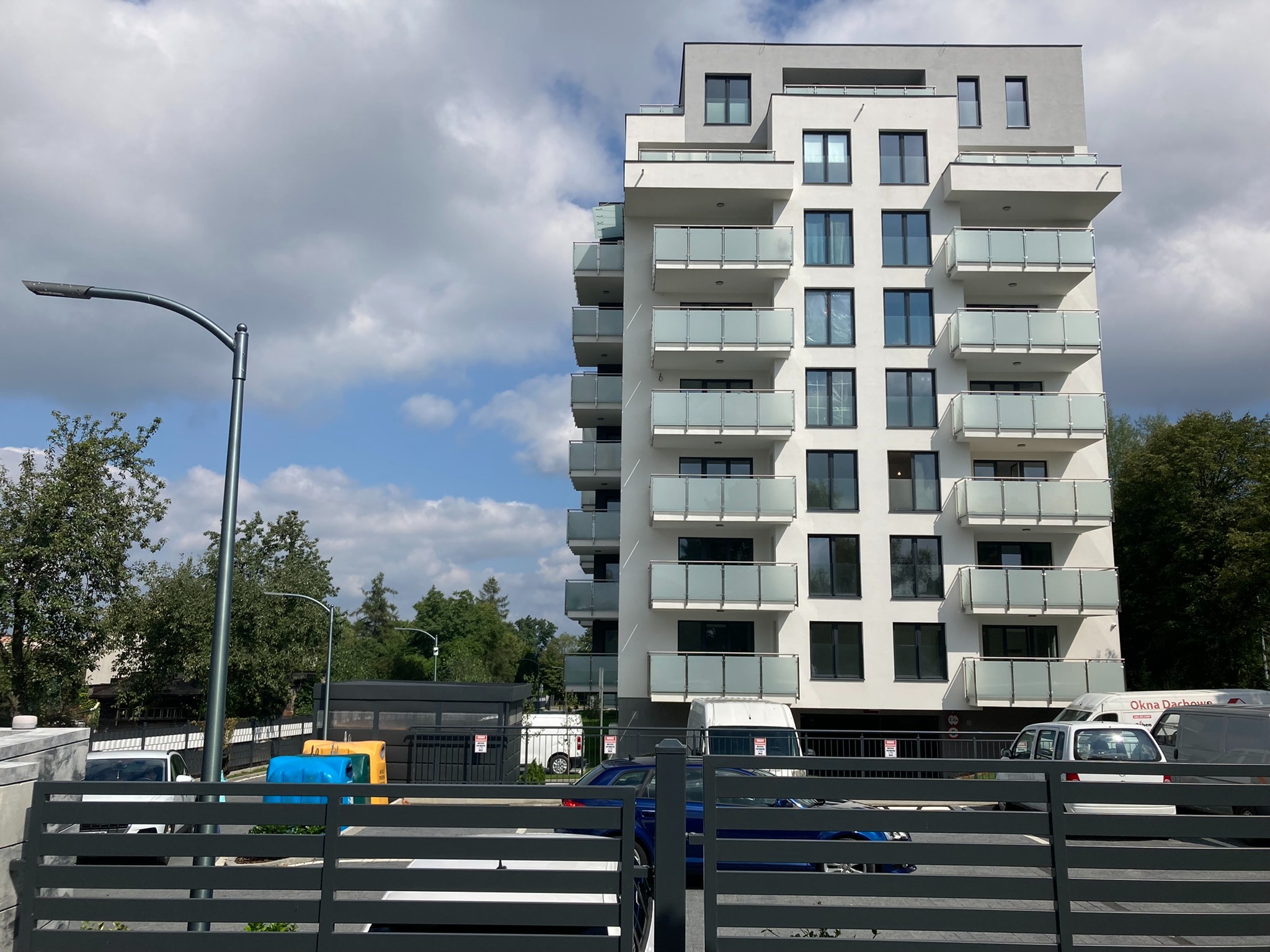 Nowoczesny, biały budynek mieszkalny z balkonami o szklanych balustradach, widziany zza szarego ogrodzenia panelowego, z zaparkowanymi samochodami i pojemnikami na śmieci w tle.