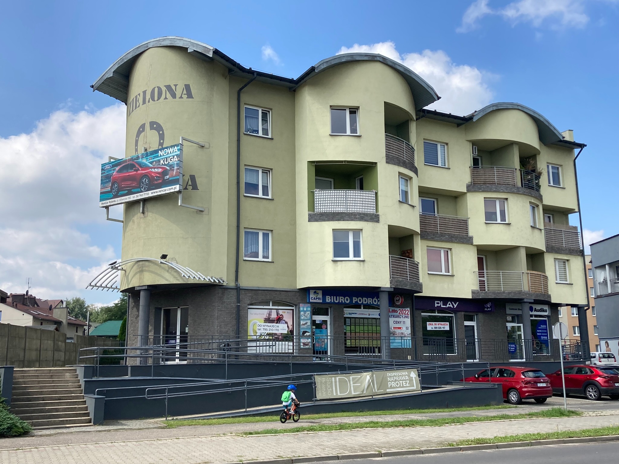 Jasnozielony budynek z zaokrąglonym narożnikiem i balkonami, na którym znajduje się reklama samochodu Nowa Kuga, na parterze lokale usługowe z biurem podróży i gabinetem protetycznym, przed...