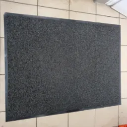 Szara wycieraczka z czarnym obramowaniem leżąca na beżowych, kwadratowych płytkach podłogowych z widocznymi zabrudzeniami i śladami użytkowania.