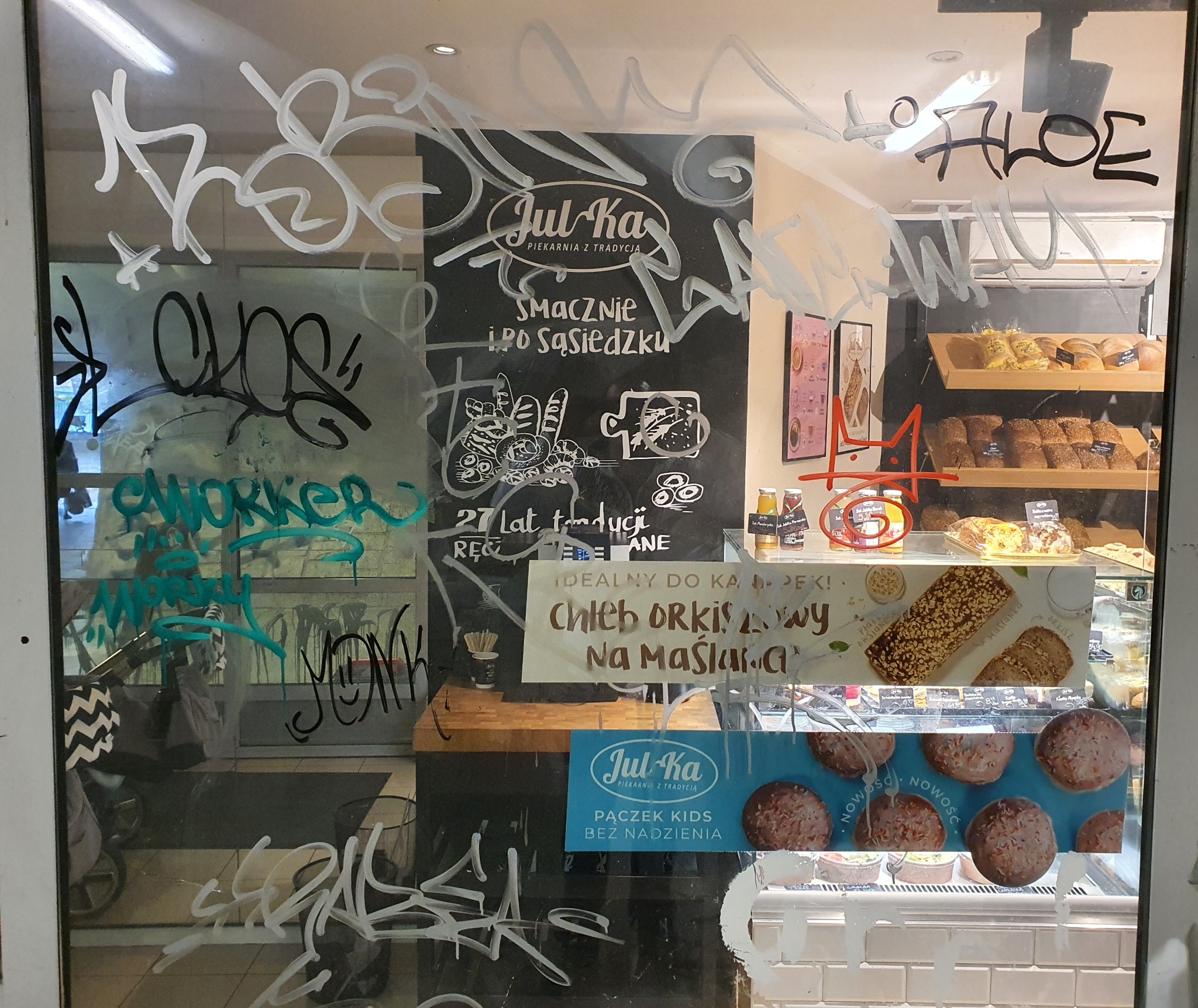 Szyba witryny piekarni z graffiti w kolorze białym i turkusowym, zasłaniająca częściowo wnętrze z półkami pełnymi pieczywa i ciast.