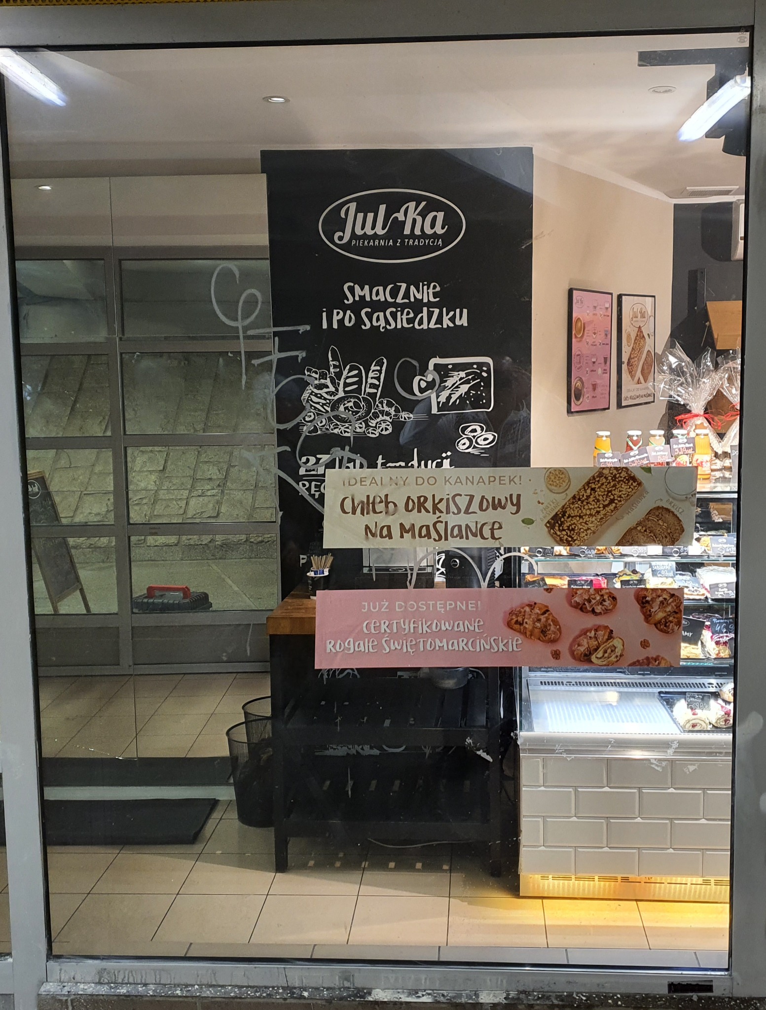 Brudna szyba piekarni Jul-Ka z rysunkami i ogłoszeniami o chlebie orkiszowym na maślance oraz certyfikowanych rogale świętomarcińskich.