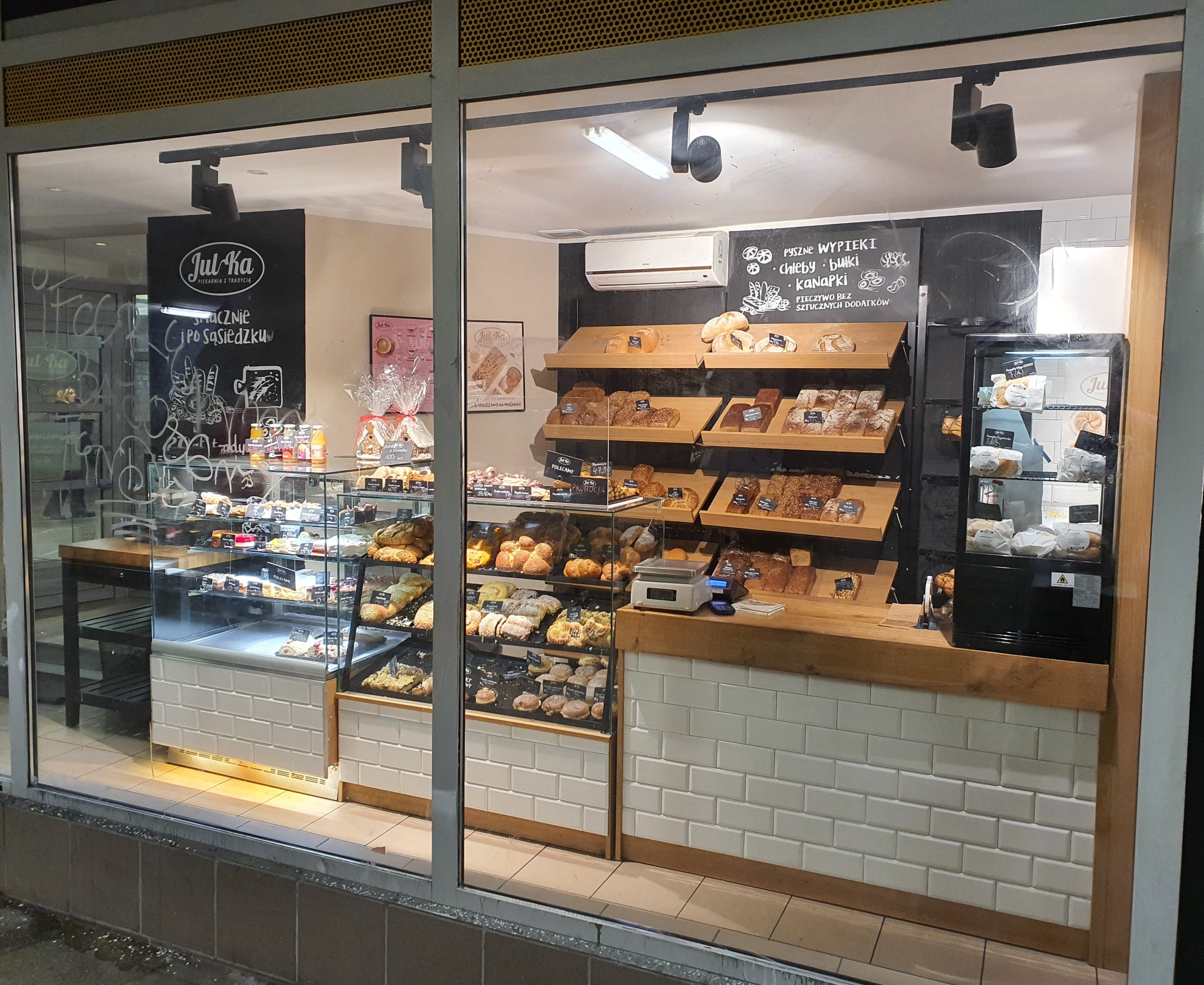 Wystawa piekarni Jul-Ka z różnorodnym pieczywem i ciastami za szybą, widoczne półki z chlebem, bułkami, ciastkami, tablica z napisem 'Pyszne wypieki' i logo piekarni.
