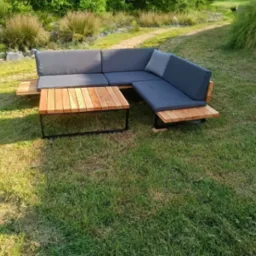 Zestaw mebli ogrodowych: narożna sofa z szarymi poduchami i drewniany stolik kawowy z metalową ramą, umieszczone na trawniku w ogrodzie.