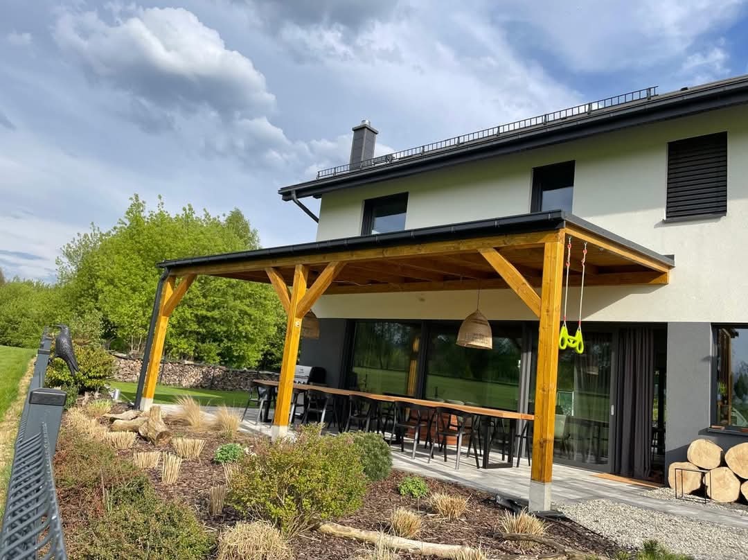 Drewniana pergola przylegająca do nowoczesnego domu z dużymi oknami, oświetlona wiszącą lampą z naturalnego materiału, z długim stołem i krzesłami na zewnątrz, otoczona starannie zaprojektowanym...