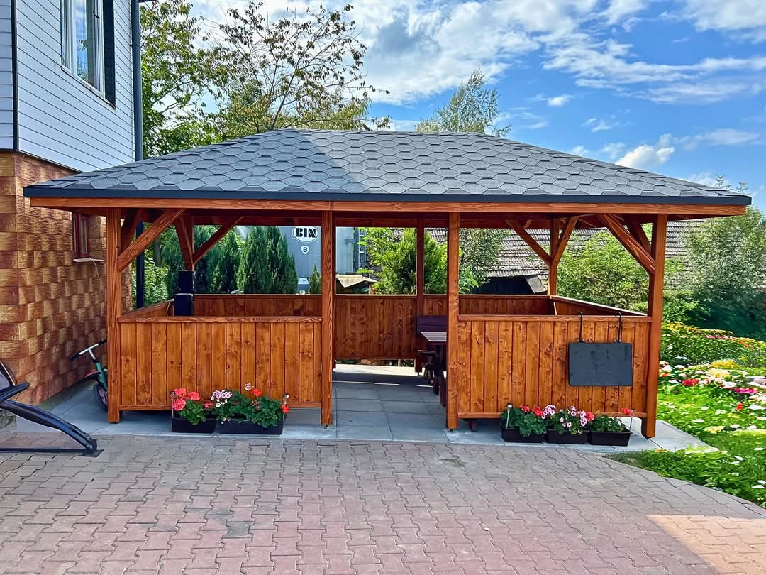 Drewniana konstrukcja z dachem z gontu bitumicznego, otoczona kwiatami w donicach, z widocznym stołem wewnątrz i elementami zabudowy na zewnątrz.