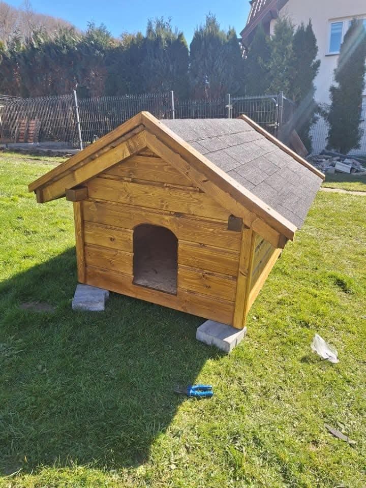 Drewniana buda dla psa z bitumicznym dachem, ustawiona na betonowych bloczkach na trawie, z nożycami do cięcia kabli leżącymi obok.