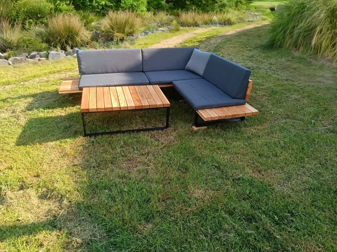 Zestaw mebli ogrodowych: narożna sofa z szarymi poduchami i drewniany stolik kawowy z metalową ramą, umieszczone na trawniku w ogrodzie.