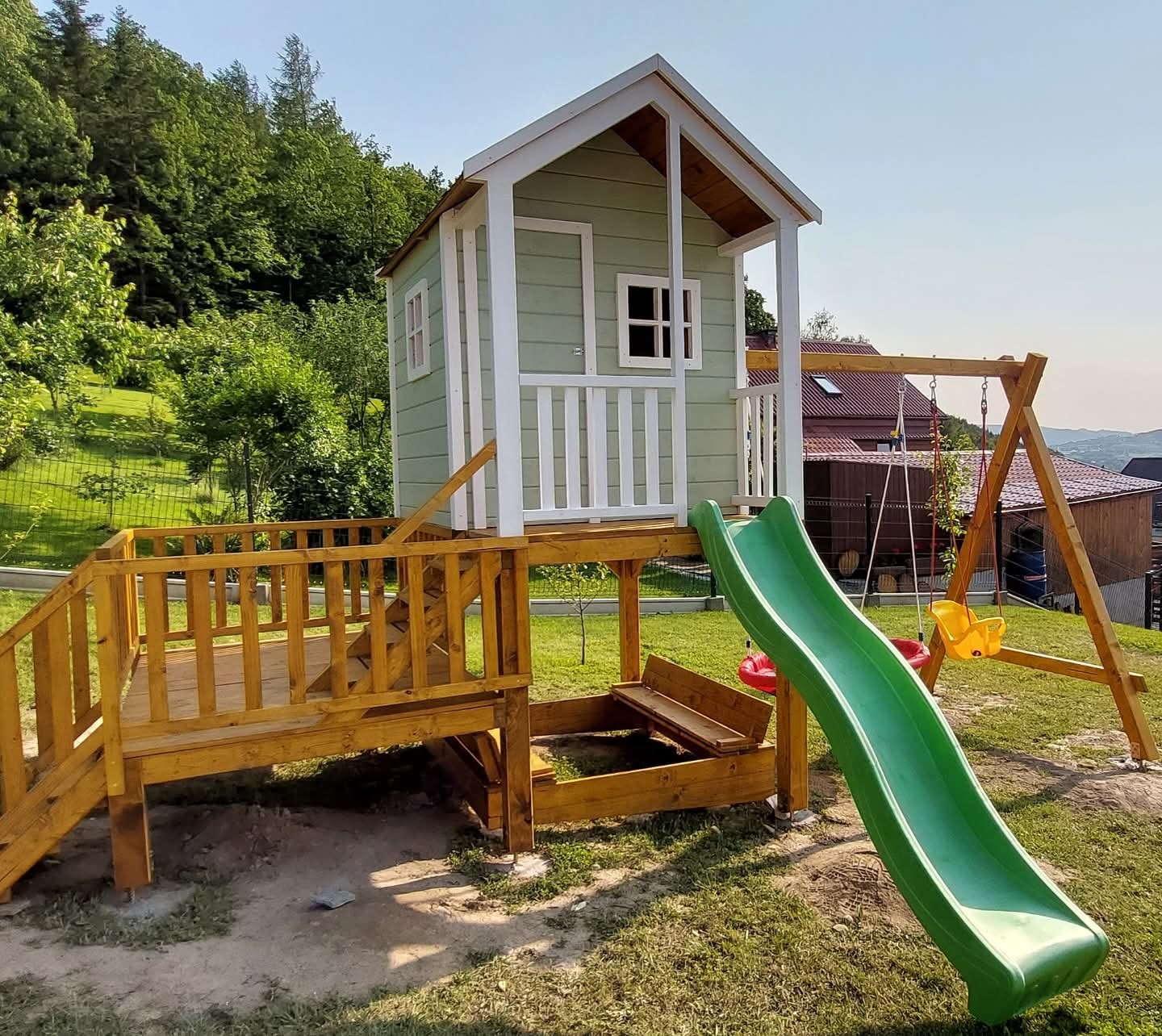 Drewniany plac zabaw z domkiem na podeście, zjeżdżalnią i huśtawką na tle zieleni. Całość w jasnych, pastelowych barwach, widok z boku, w słoneczny dzień.