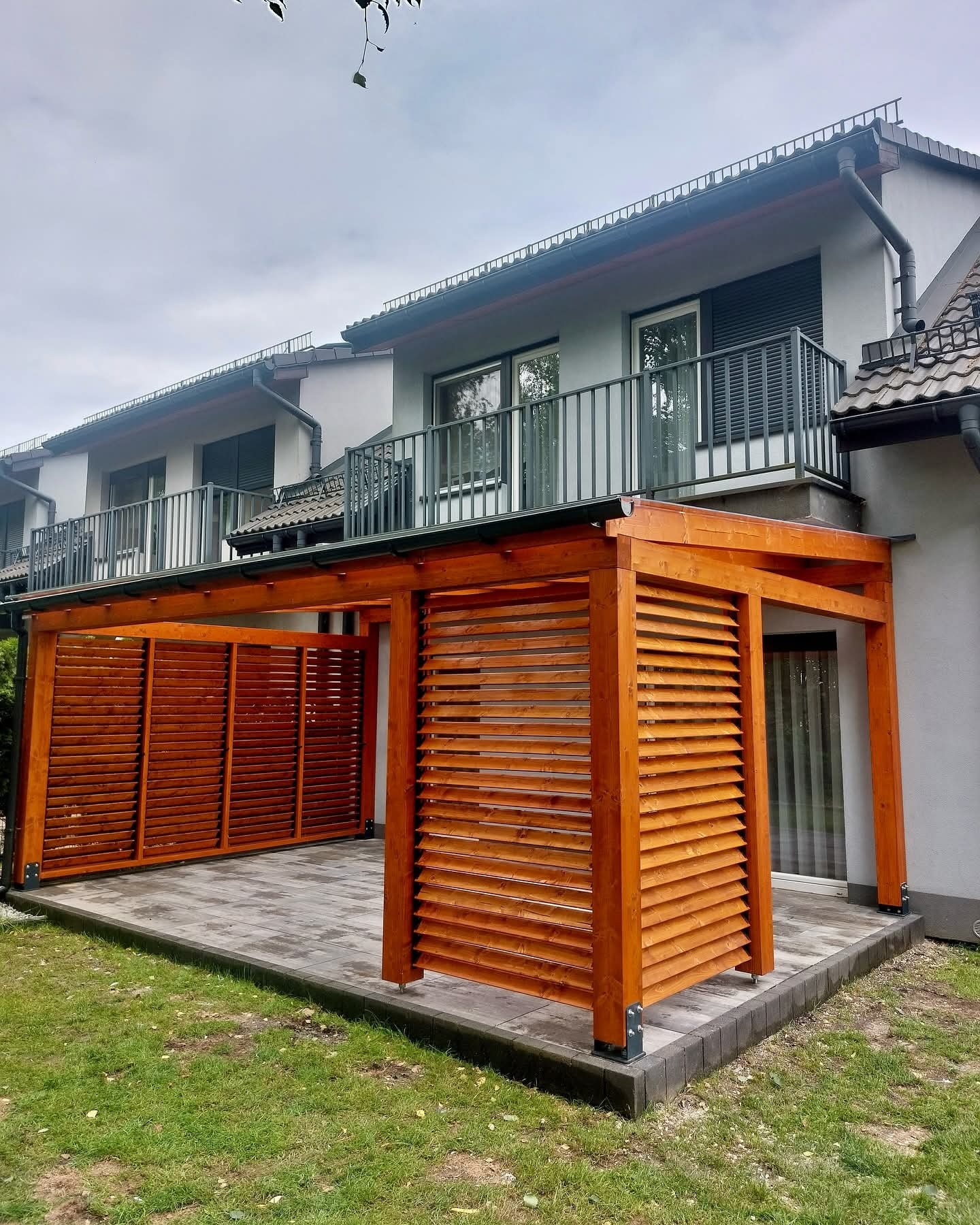 Drewniana konstrukcja z lamelami, przylegająca do budynku. Uwagę przyciągają metalowe łączniki u podstawy słupów i precyzyjnie ułożona kostka brukowa. Całość w naturalnej kolorystyce.