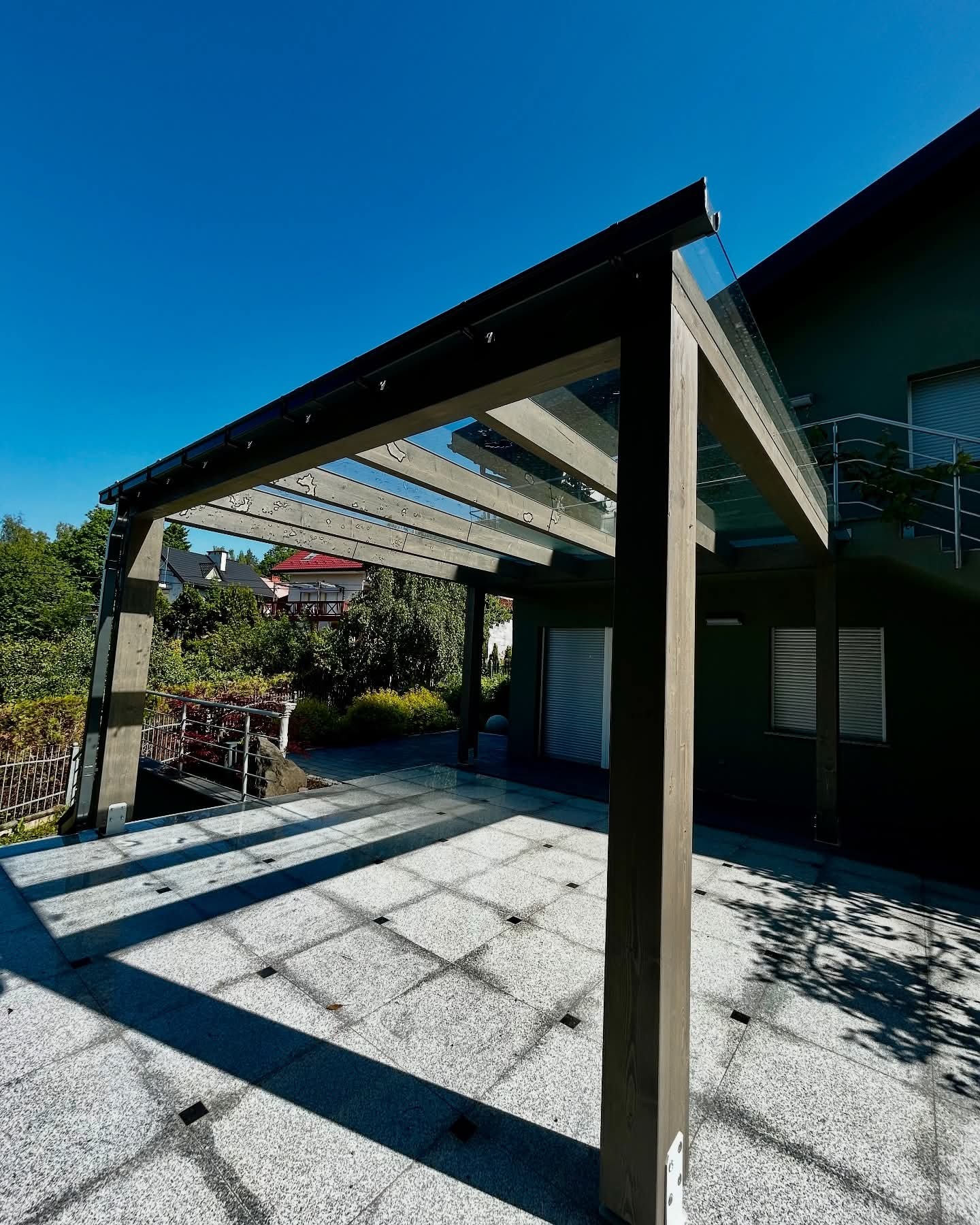 Nowoczesna pergola z drewnianą konstrukcją i szklanym dachem, przylegająca do budynku z zieloną elewacją. Cień rzucany przez konstrukcję na jasną, kwadratową posadzkę.