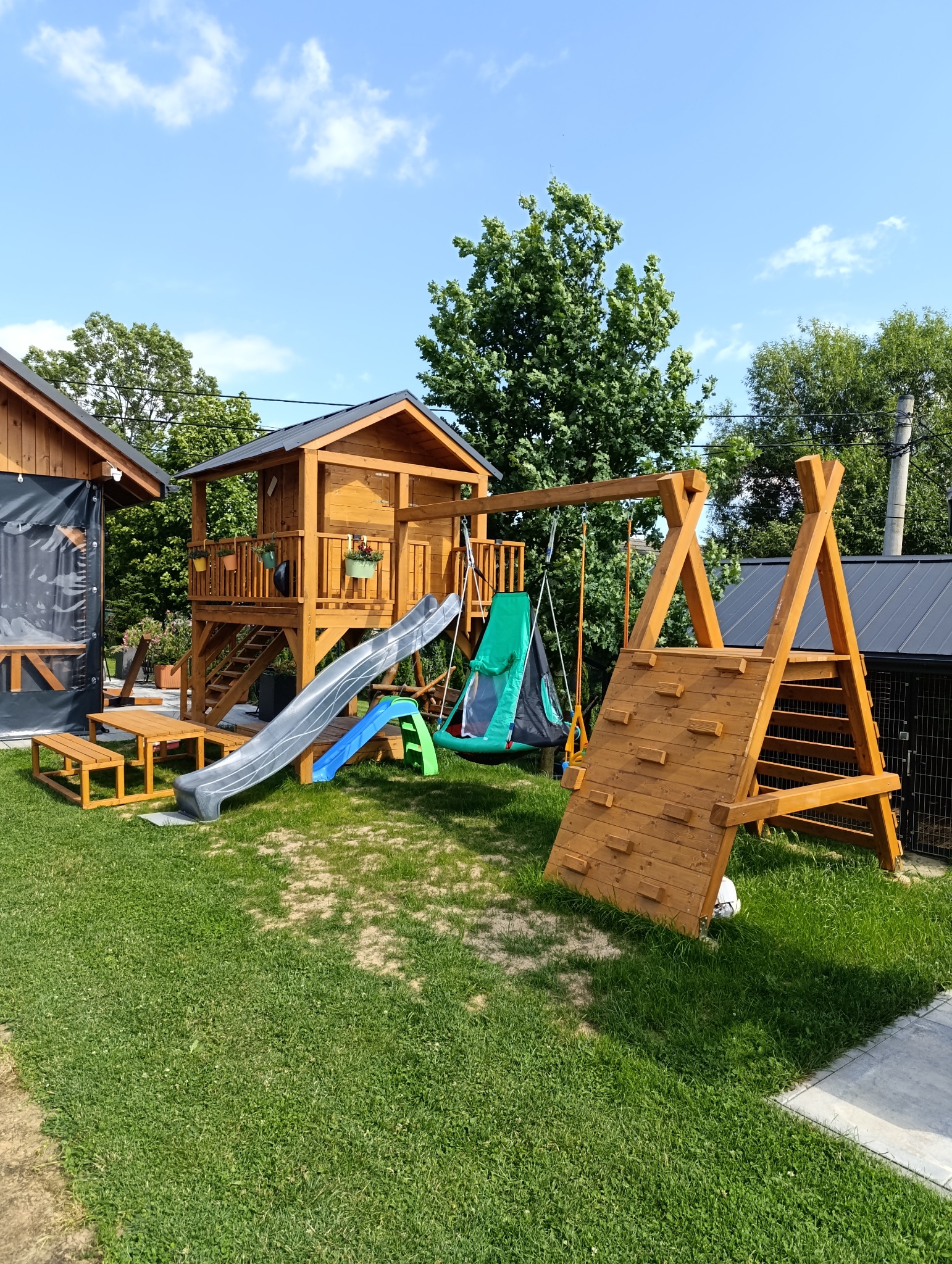 Drewniany plac zabaw z domkiem na palach, zjeżdżalnią, huśtawką i ścianką wspinaczkową na trawniku w słoneczny dzień. Widoczny stół piknikowy i budynek w tle.