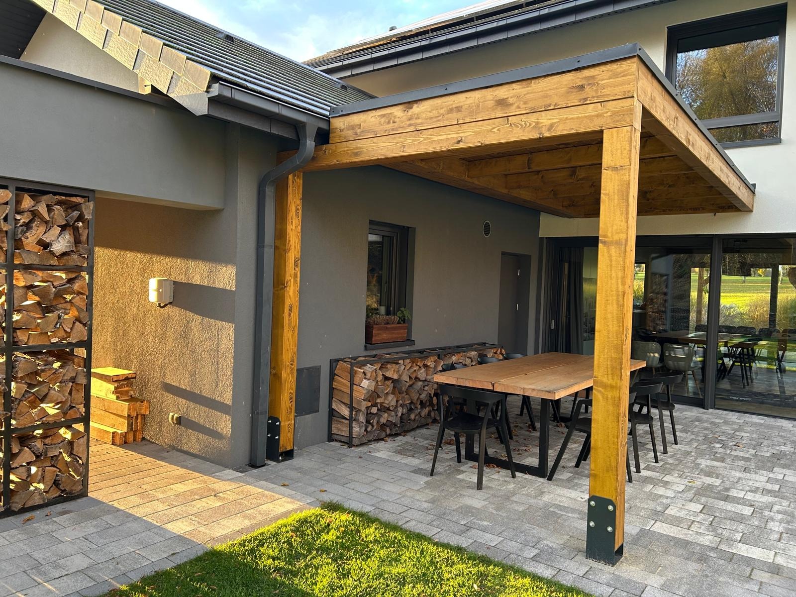 Drewniana pergola z solidnymi słupami, przylegająca do domu z szarą elewacją. Na tarasie stół z krzesłami i dekoracyjne miejsce na drewno. Widok na ogród.