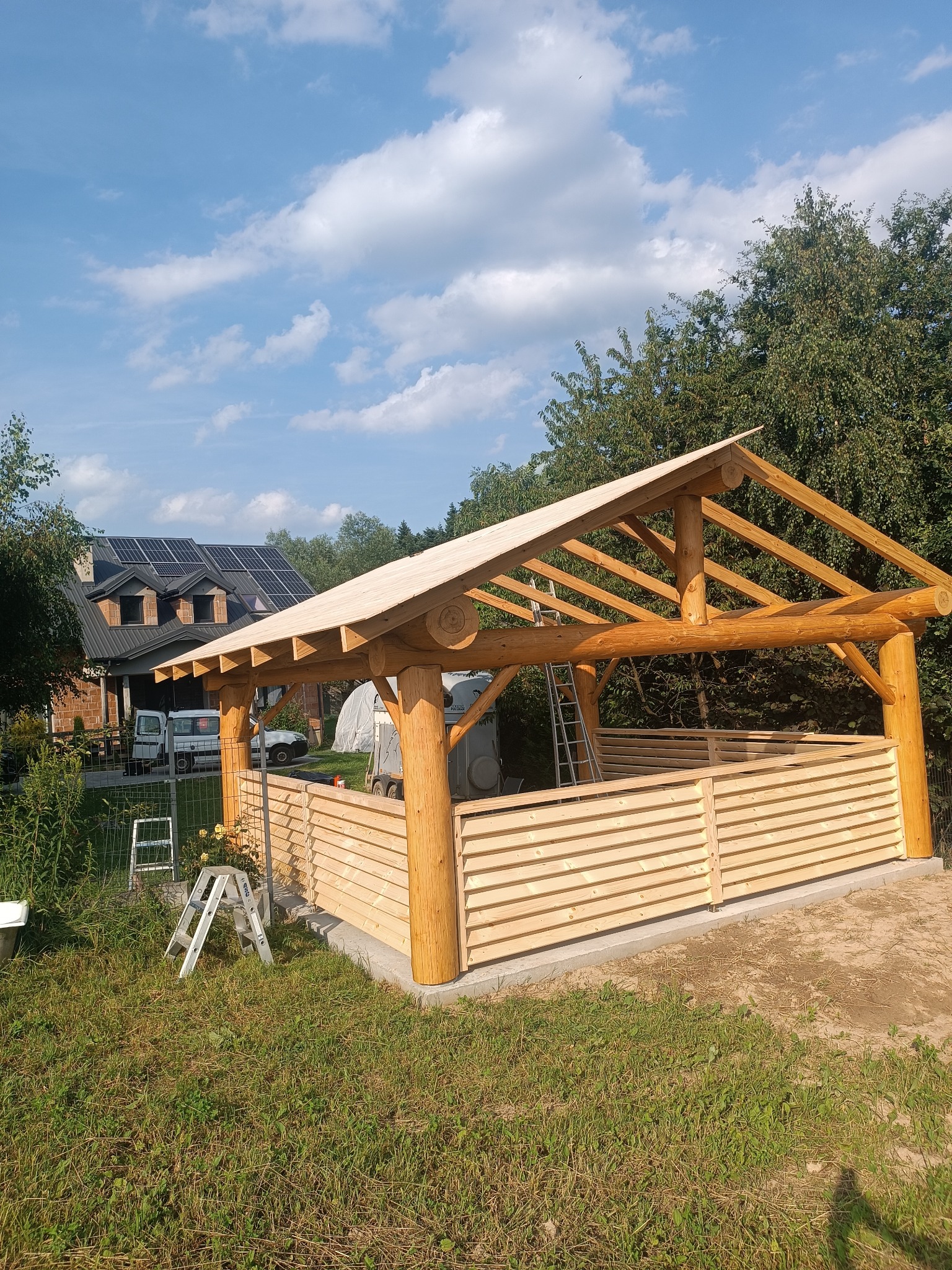 Konstrukcja drewnianej altany z bali w trakcie budowy, widoczny szkielet dachu i częściowe wypełnienie ścian lamelami, drabina oparta o konstrukcję, dom z panelami słonecznymi w tle.