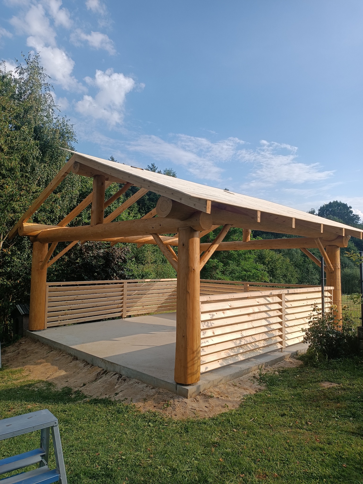 Surowa konstrukcja z bali, prawdopodobnie altany, z drewnianym wypełnieniem ścian, na betonowej wylewce, w otoczeniu zieleni i błękitnego nieba.