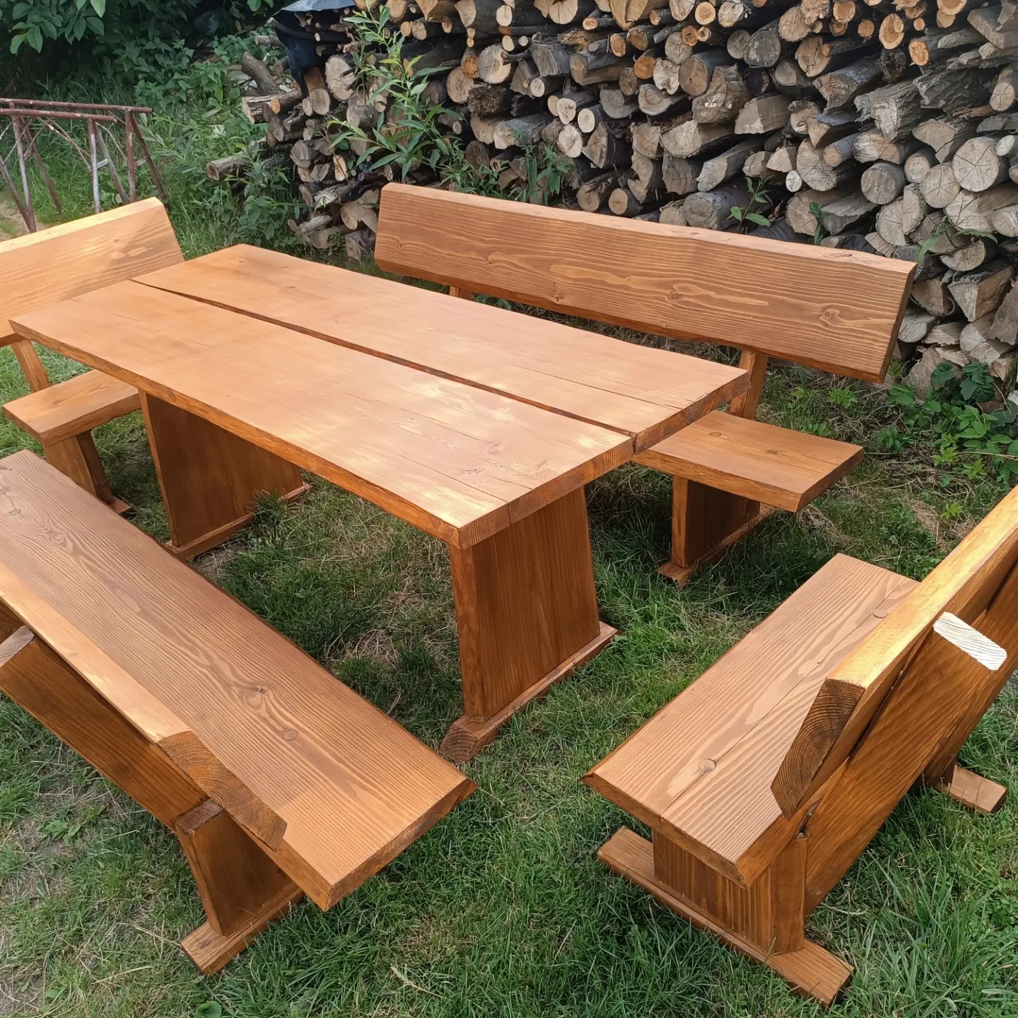 Rustykalny zestaw mebli ogrodowych z litego drewna: stół i cztery ławki z oparciami, ustawione na trawie, z ułożonymi polanami drewna w tle.