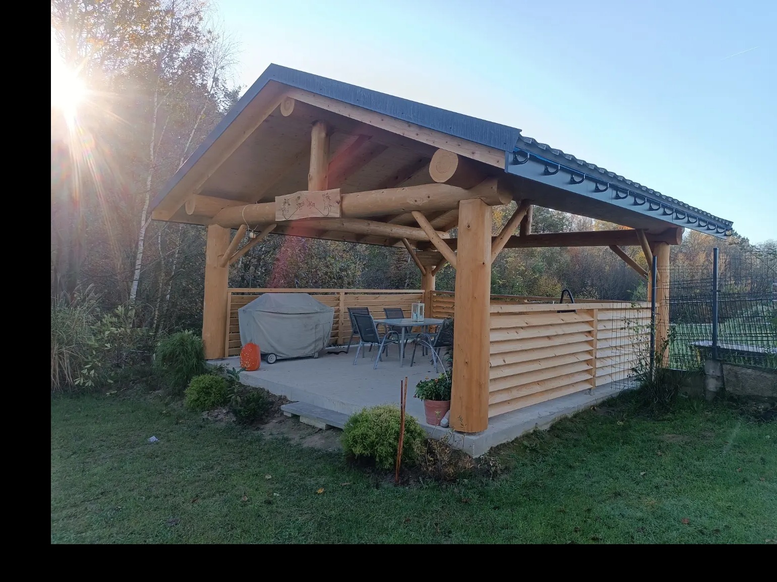 Drewniana konstrukcja z zadaszeniem, prawdopodobnie altana, z drewnianym ażurowym panelem bocznym, betonową podłogą, meblami ogrodowymi pod spodem i grillem przykrytym pokrowcem, otoczona zielenią...