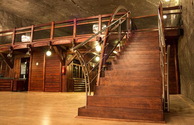 Wnętrze z drewnianymi schodami i balustradą ze stali nierdzewnej. Ciepłe oświetlenie i drewniane wykończenia tworzą przytulną atmosferę.