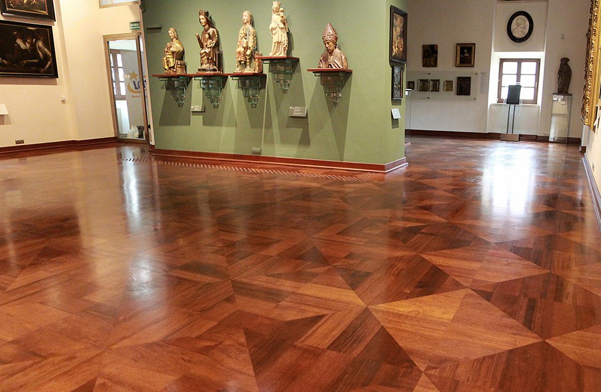 Parkiet z egzotycznego drewna ułożony w geometryczny wzór gwiazdy, w muzeum z ekspozycją rzeźb i obrazów. Oświetlenie dzienne podkreśla głębię kolorów drewna.