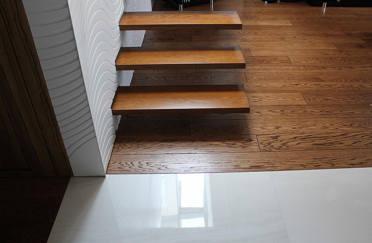 Nowoczesne schody z drewna dębowego, wspornikowe, z subtelnym oświetleniem. Parkiet dębowy w ciepłym odcieniu, kontrastujący z białą, falistą ścianą i jasnymi płytkami.