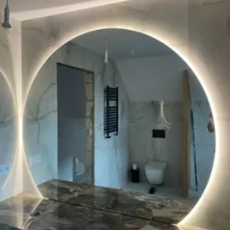 Okrągłe lustro z podświetleniem LED w łazience w trakcie remontu, z widoczną marmurową umywalką, fragmentem instalacji grzewczej i niedokończonym wejściem do drugiego pomieszczenia.