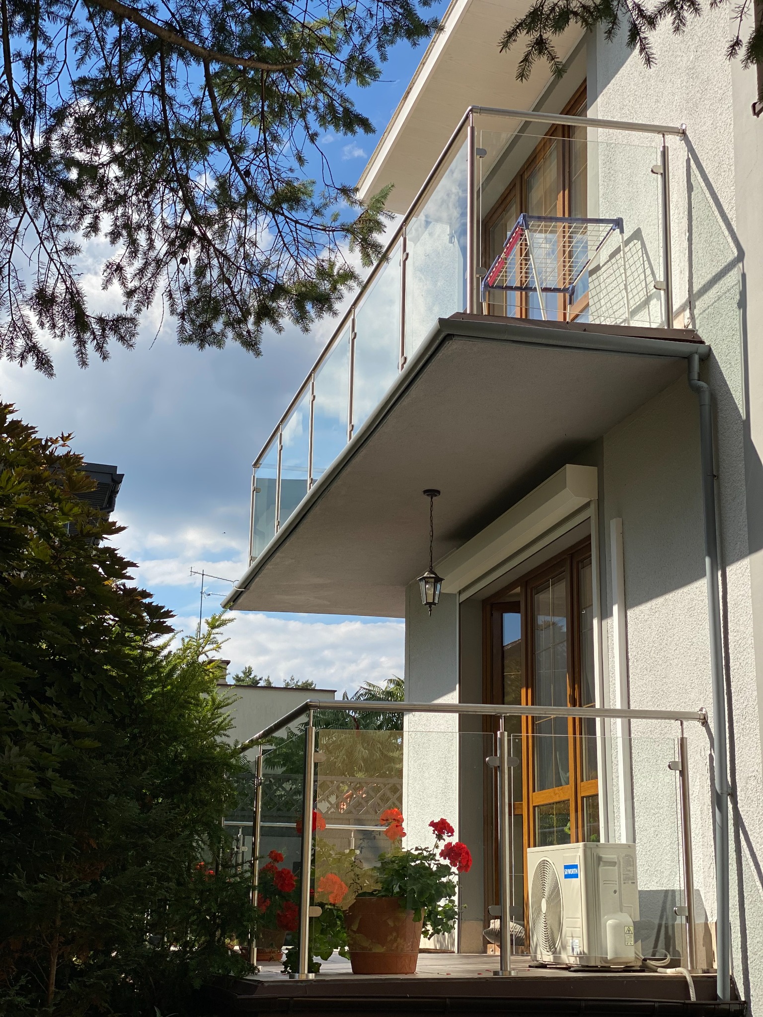Nowoczesny balkon z balustradą szklaną i metalowymi elementami, widoczne kwiaty w donicach i klimatyzator na dolnym balkonie, suszarka na pranie na górnym balkonie, w tle błękitne niebo z chmurami.