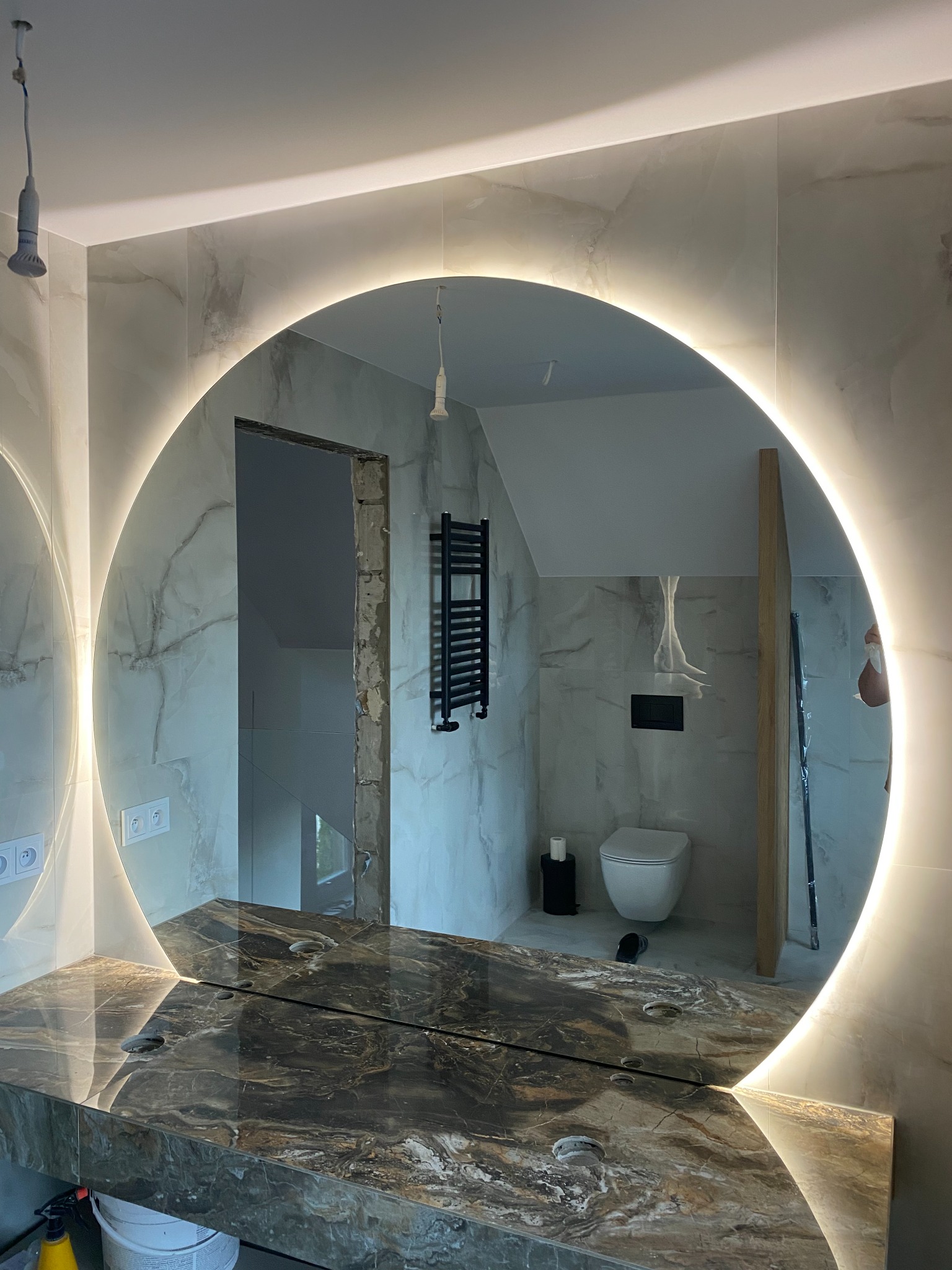 Okrągłe lustro z podświetleniem LED w łazience w trakcie remontu, z widoczną marmurową umywalką, fragmentem instalacji grzewczej i niedokończonym wejściem do drugiego pomieszczenia.
