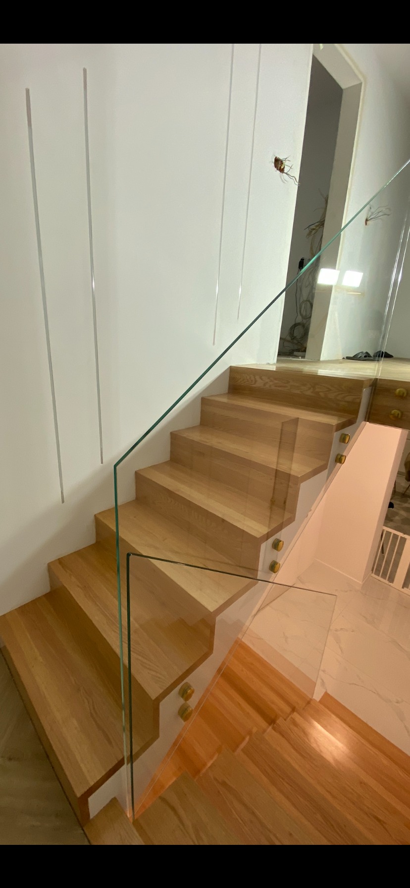 Nowoczesne schody z drewnianymi stopniami i szklaną balustradą mocowaną na złotych uchwytach. Widoczne niedokończone prace elektryczne na ścianie.