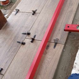 Patryk Tchórz - Układanie płytek imitujących drewno na zewnątrz, z użyciem poziomicy, systemu poziomowania i szlifierki kątowej w tle.