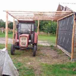 Patryk Tchórz - Konstrukcja z surowego drewna nad traktorem, prawdopodobnie wiata lub zadaszenie w trakcie budowy, z widocznym ociepleniem na ścianie budynku w tle.