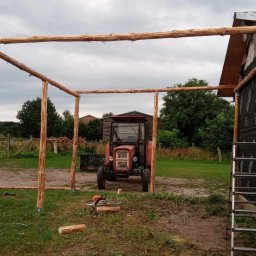 Patryk Tchórz - Konstrukcja z surowego drewna, prawdopodobnie zadaszenie lub wiata, w tle traktor i budynek gospodarczy, teren budowy z piłą łańcuchową.