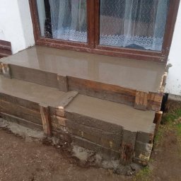 Patryk Tchórz - Wylewanie betonowych schodów wejściowych do domu z wykorzystaniem drewnianego szalunku, widoczna łopata oparta o ścianę budynku.