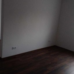 Patryk Tchórz - Pusty pokój z ciemną podłogą, świeżo pomalowanymi szarymi ścianami i oknem z widocznymi akcesoriami na parapecie.