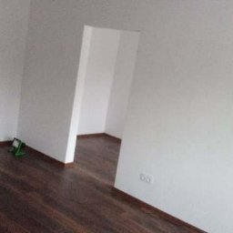 Patryk Tchórz - Pomalowane na biało wnętrze pokoju z ciemną podłogą, widoczne gniazdko elektryczne i zielony poziomica laserowa przy ścianie.