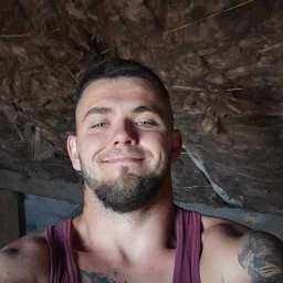 Patryk Tchórz - Uśmiechnięty mężczyzna w podkoszulku na tle surowego, brązowego stropu z widocznymi mocowaniami.